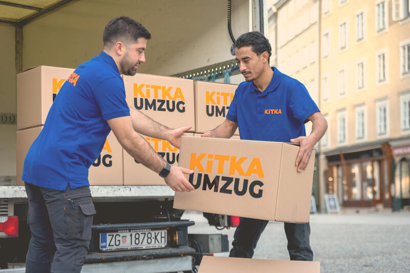 Zwei Umzugshelfer von KITKA heben einen Umzugskarton in der Salzburger Altstadt